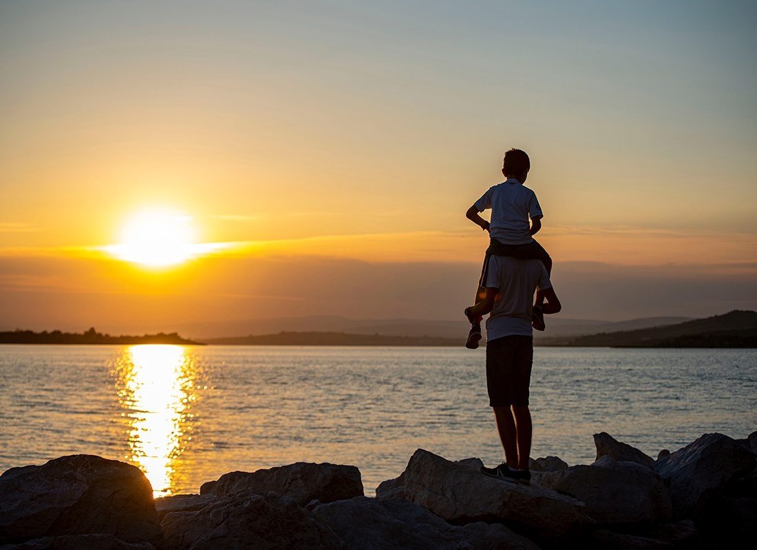 About Our Agency - Rear View of a Father Holding His Son on His Shoulders as They Both Watch the Sunset Over the Water