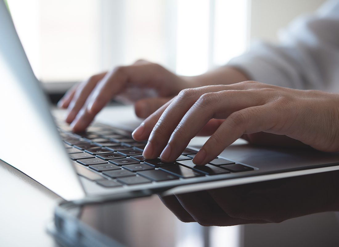 Blog - Close Up of Professional Typing on Open Laptop on a Glass Desk Next to a Window